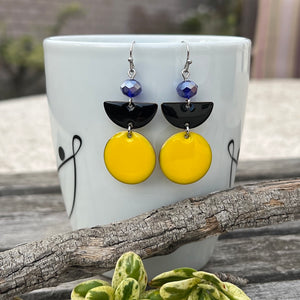 These beautiful geometric earrings feature a double-sided copper circle and semi-circle with an oil drip enamel finish in yellow and black. A cobalt blue faceted crystal rondelle with an aurora borealis finish completes the look. Earrings measure approximately 2" long on stainless steel ear wires with silicone backs.